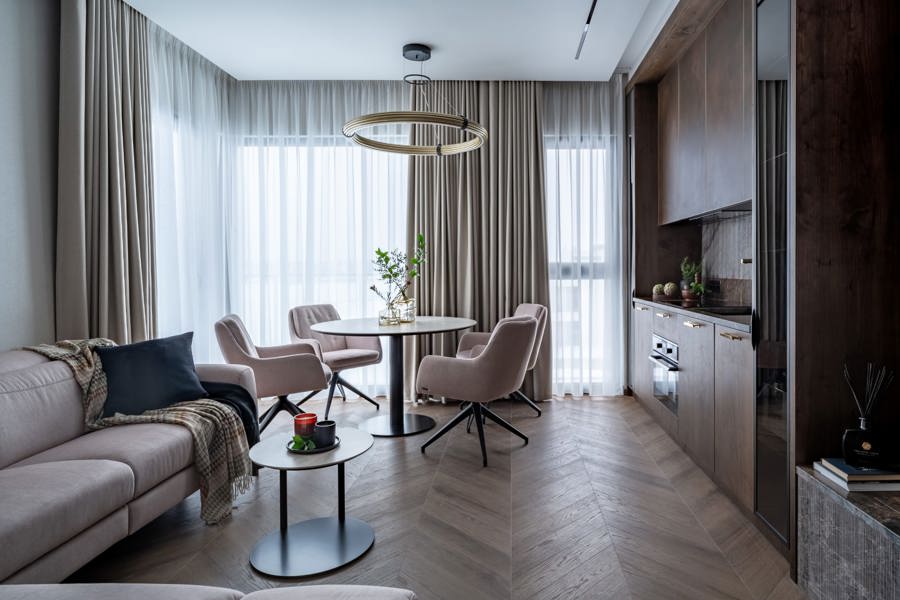 Otwarty salon połączony z jadalnią i aneksem kuchennym — beżowa sofa, okrągły stół z czterema pudrowymi fotelami i duże okno z zasłonami.